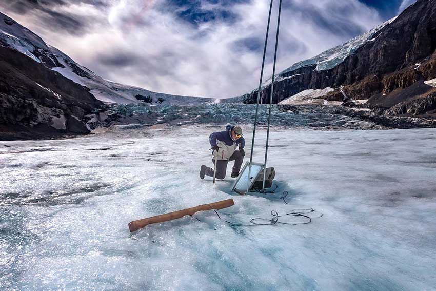 Researcher is kneeling on the Athabasca Glacier looking at a monitoring station sitting on the ice