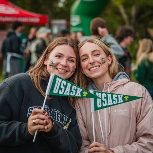 Two women in USask clothing pose with USask pennants in their hands.